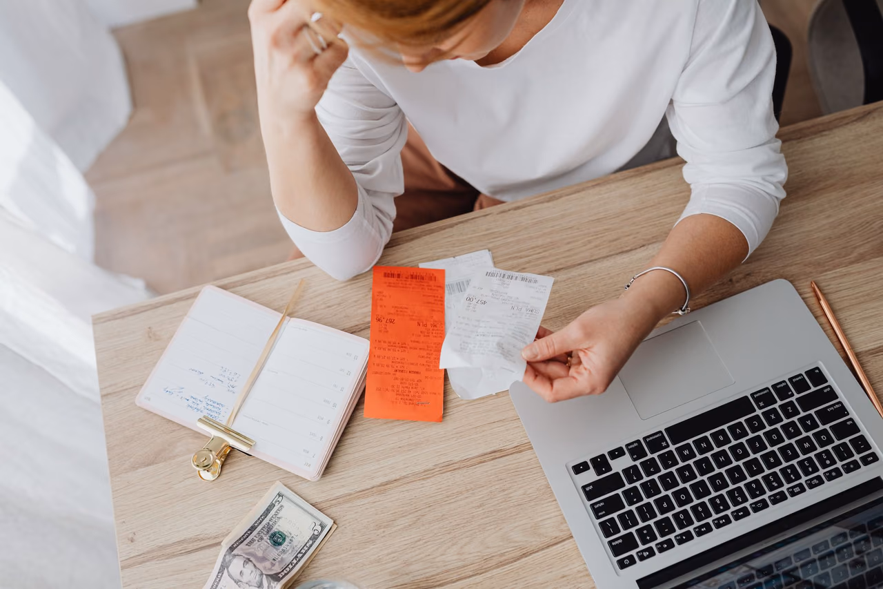 A person at a wooden desk holding a paper receipt, with an open laptop, a notebook, a pen and a few dollar bills nearby