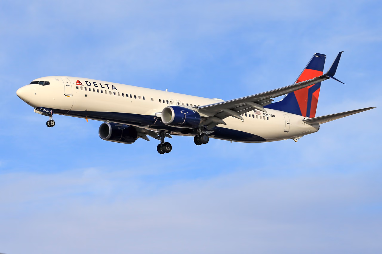 A white and navy Delta Boeing 737 with a red and blue tail, landing gear down, flying against a blue sky with thin clouds.