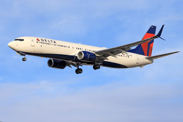 A white and navy Delta Boeing 737 with a red and blue tail, landing gear down, flying against a blue sky with thin clouds.