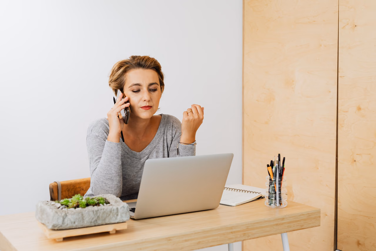 A woman on a phone call at a wooden desk with an open laptop, a notebook and a planter of succulents.