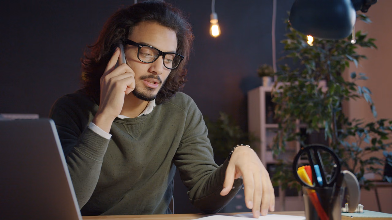 A man in glasses talks on the phone at his desk beside a laptop, reading over papers in a dimly lit home office.