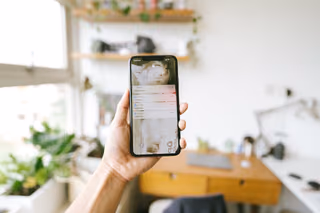 A hand holds up a phone showing a stack of unread notifications on its lock screen, in a sunny home office with plants and a wooden desk