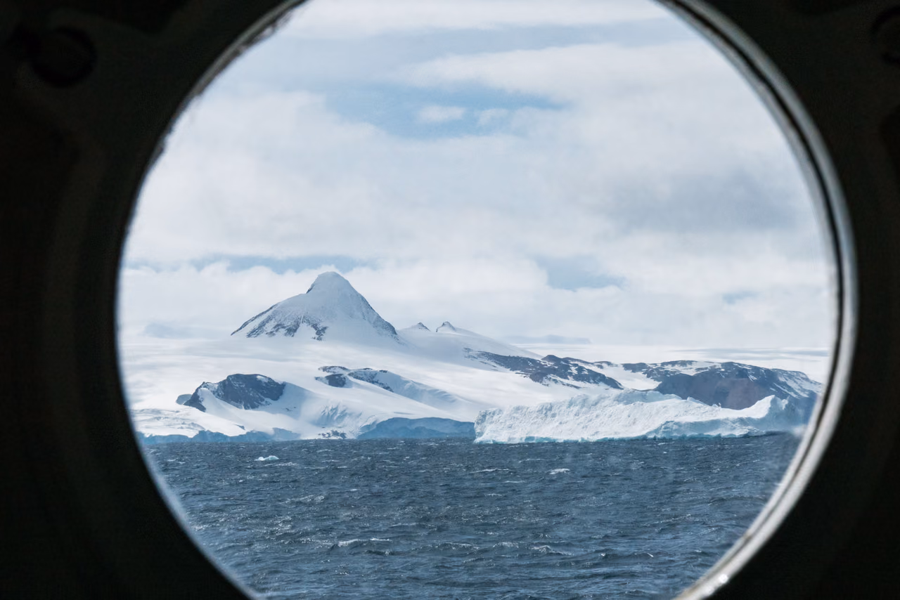 A snow-covered peak and glacier-draped coastline above a choppy gray sea, framed by a ship's round porthole.