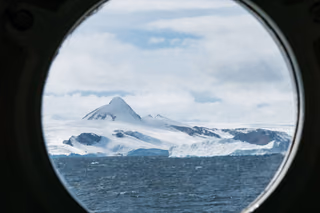 A snow-covered peak and glacier-draped coastline above a choppy gray sea, framed by a ship's round porthole.