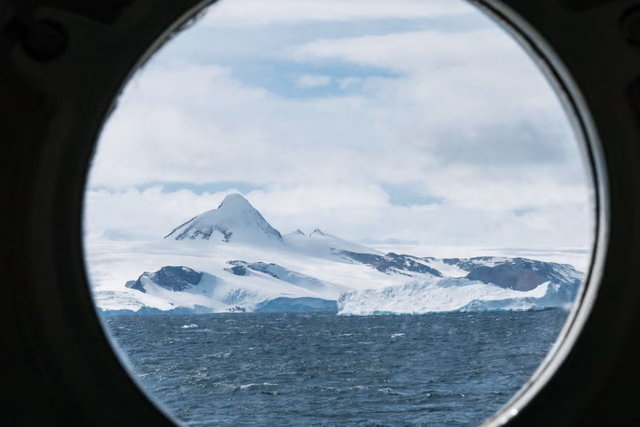 A snow-covered peak and glacier-draped coastline above a choppy gray sea, framed by a ship's round porthole.