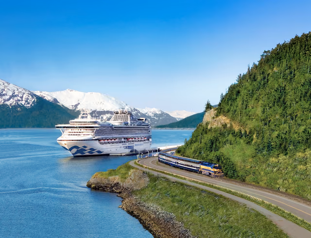 A Princess cruise ship docked beside a blue and yellow train on a shoreline track, with snow-capped mountains behind