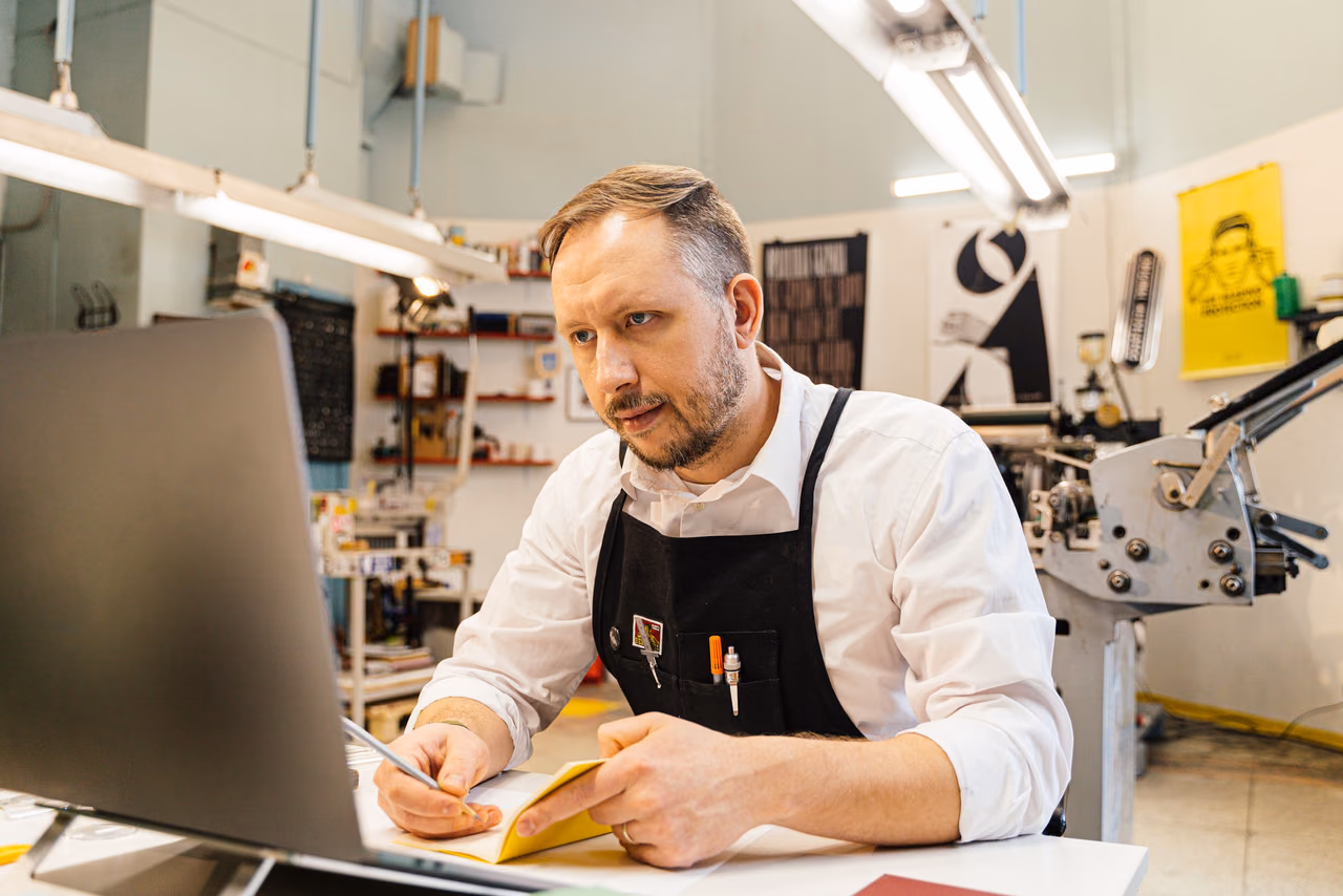A shop owner in a white shirt and black apron takes notes while looking at a laptop in his print shop, with a printing press and posters behind him.