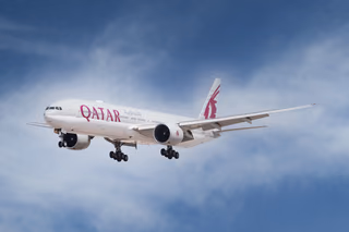 A Qatar Airways Boeing 777 on approach with its landing gear down, white fuselage and burgundy oryx tail against a blue sky with thin cloud