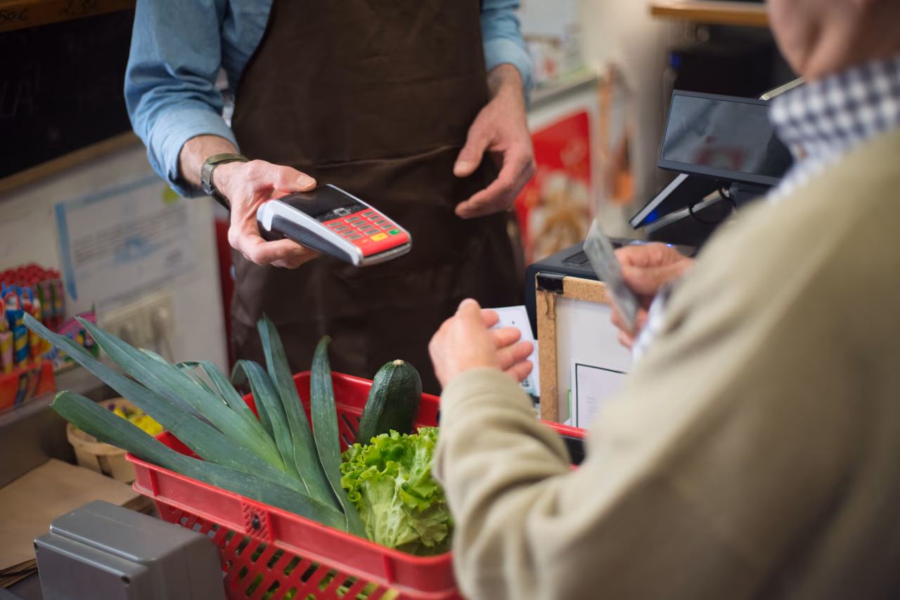 A shopper holds out a card to pay while a cashier offers a card reader over a red basket of leeks, lettuce and a zucchini