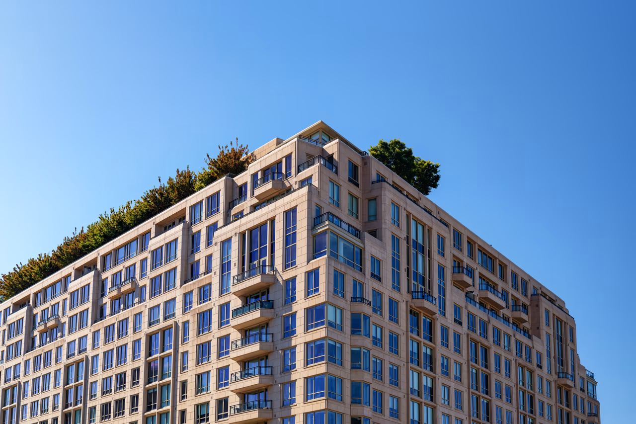 The upper storeys of a modern apartment building with balconies and rooftop trees against a clear blue sky