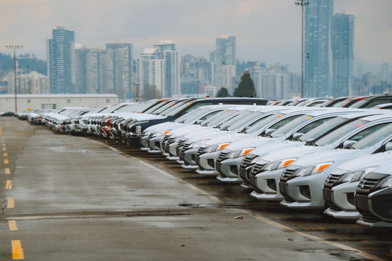 Rows of white and silver cars parked nose to nose in a wet outdoor lot, with high-rise towers behind them