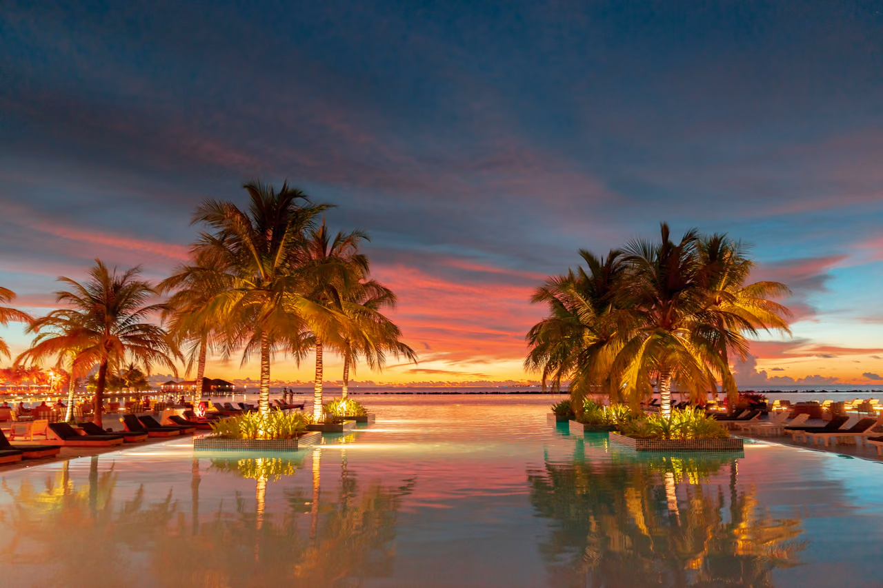 An infinity pool at a beachfront resort at dusk, with lit palm trees and sun loungers reflected in the water under a pink and blue sky.