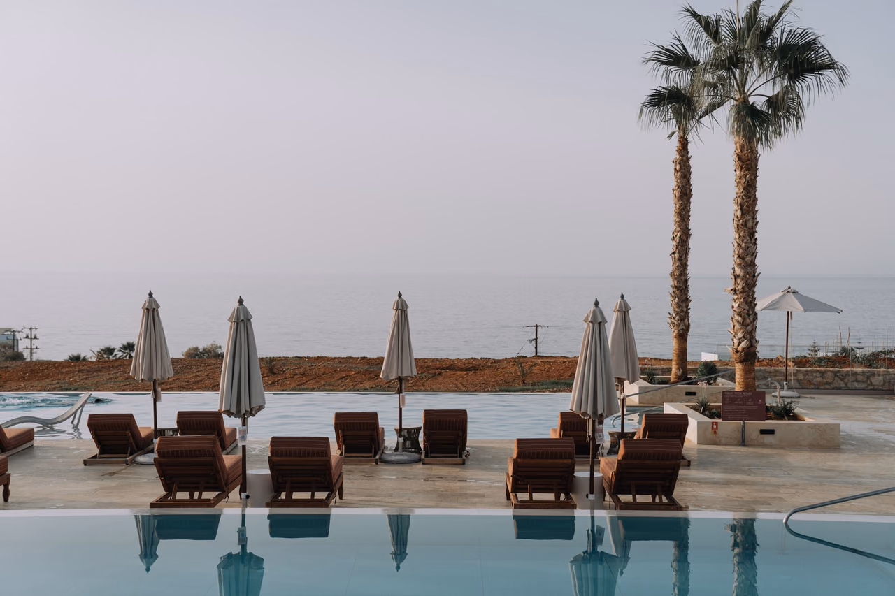 Rows of brown lounge chairs and closed cream umbrellas beside an infinity pool at a resort, with two palm trees and the sea beyond