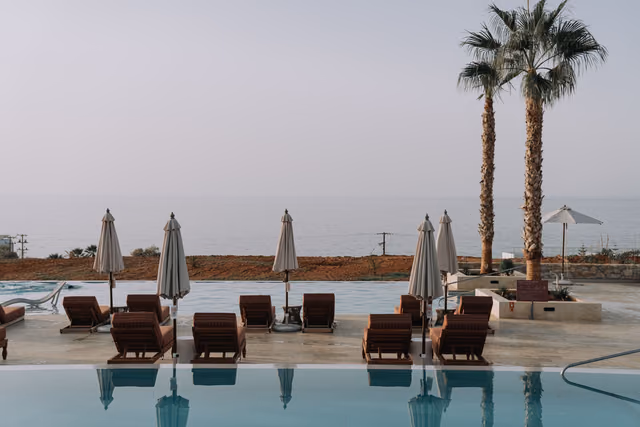 Rows of brown lounge chairs and closed cream umbrellas beside an infinity pool at a resort, with two palm trees and the sea beyond