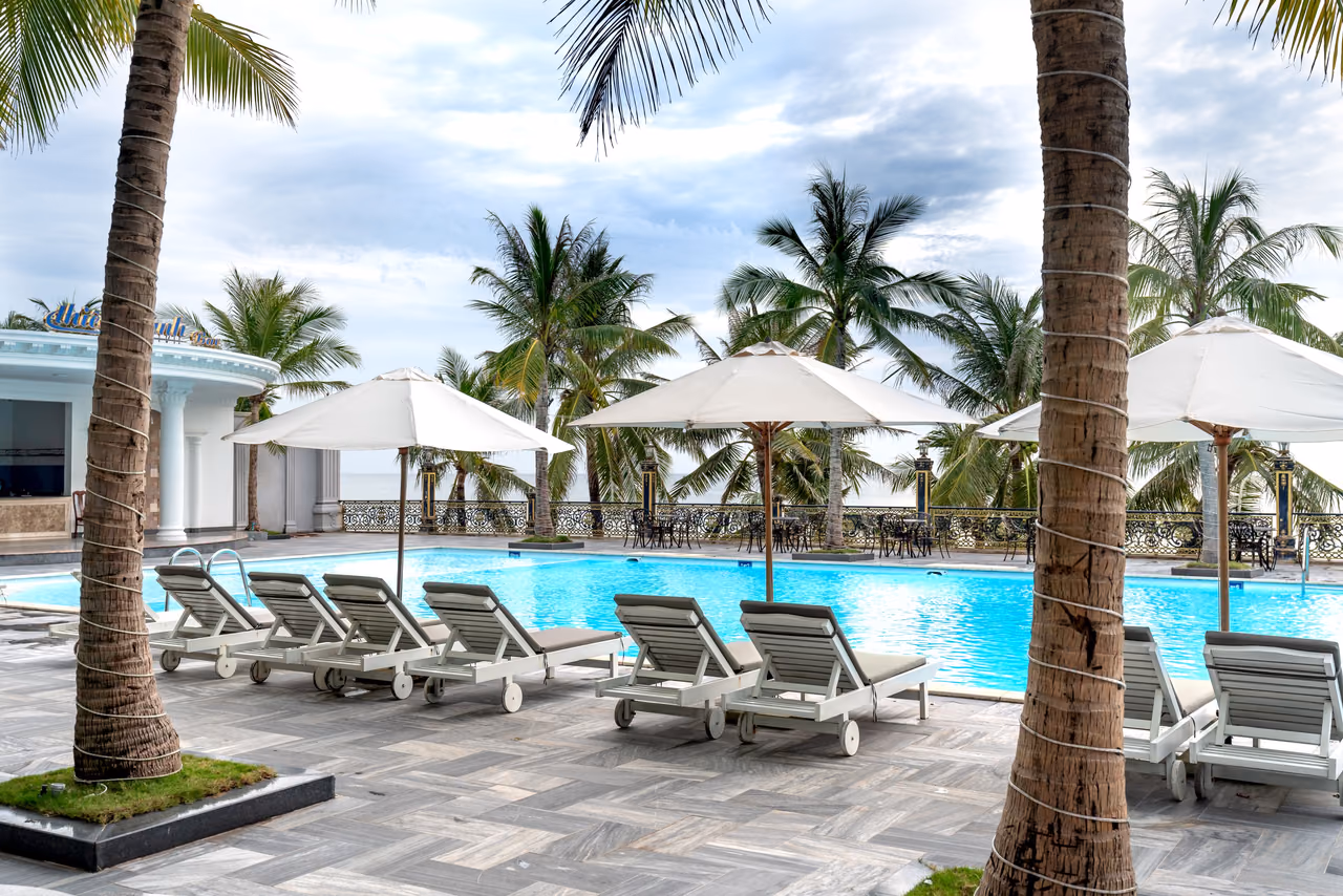 A resort swimming pool lined with white lounge chairs and patio umbrellas, framed by tall palm trees under a partly cloudy sky.