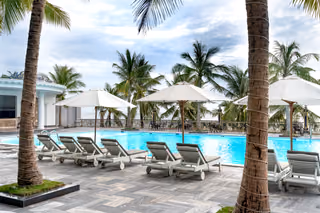 A resort swimming pool lined with white lounge chairs and patio umbrellas, framed by tall palm trees under a partly cloudy sky.