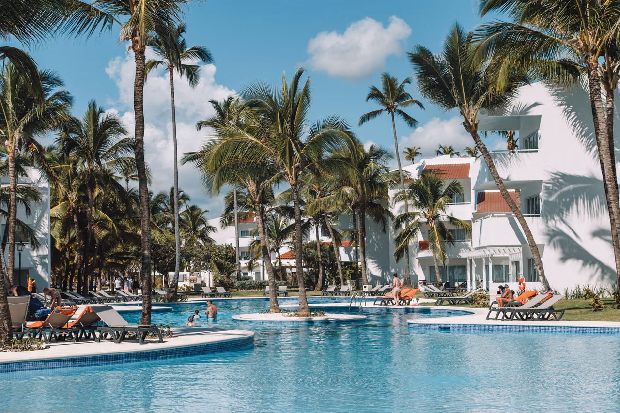 A resort pool ringed by palm trees and sun loungers in front of white hotel buildings under a blue sky.