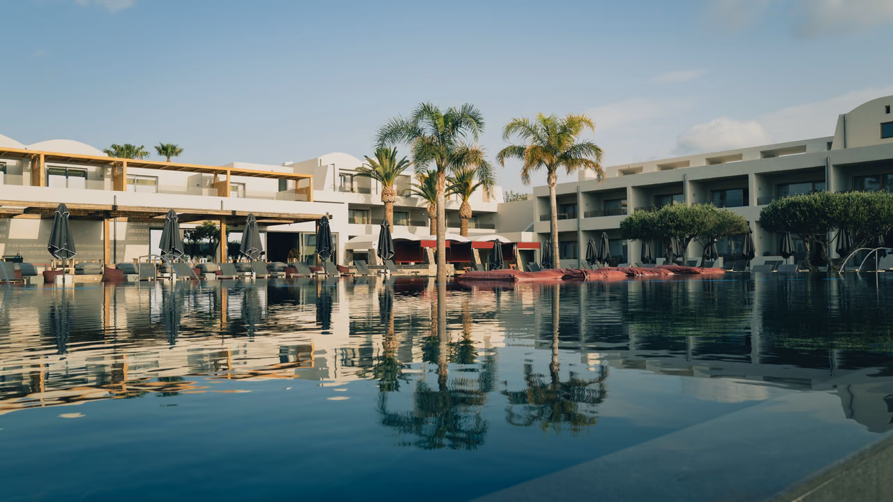 Resort pool reflecting palm trees and low white hotel buildings, with red loungers along the far edge under a pale sky