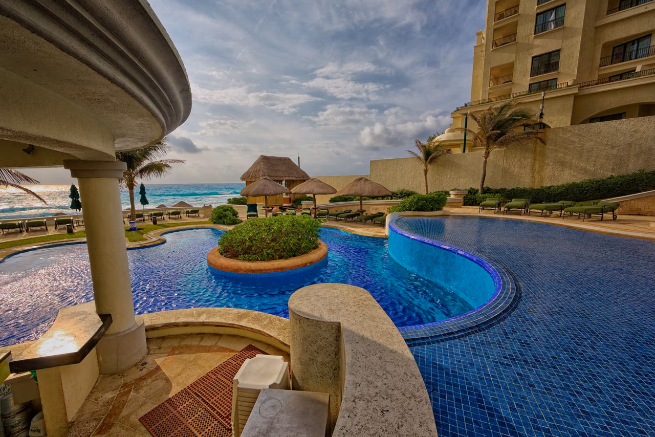 A resort swimming pool with sun loungers and palm trees beside the ocean in Mexico