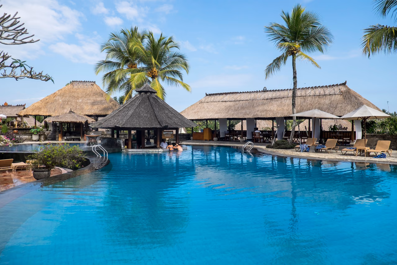A blue resort swimming pool framed by palm trees and thatched-roof pavilions under a clear sky.