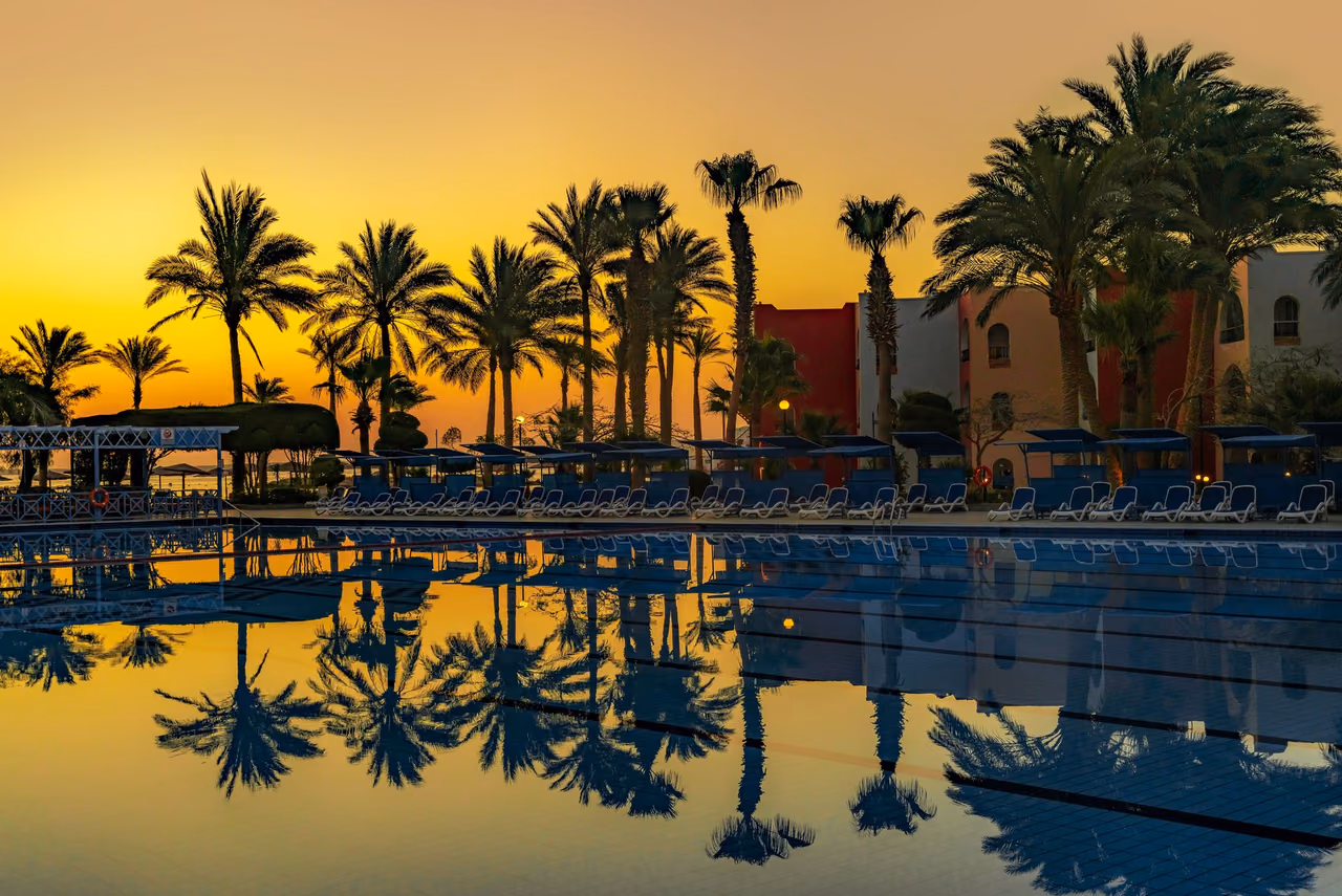A resort swimming pool at sunset, with palm trees and rows of sun loungers reflected in the still water under an orange sky