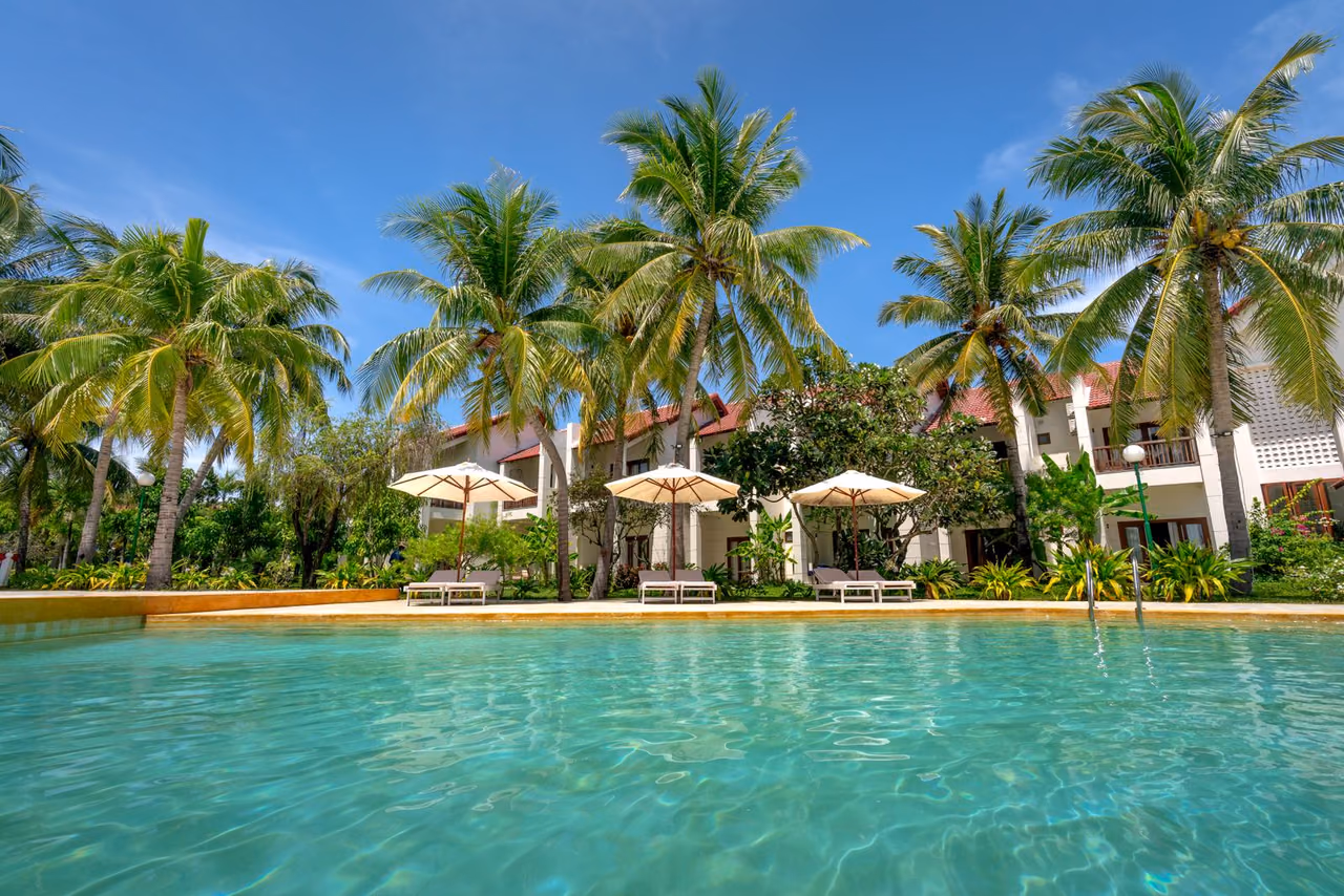 Turquoise resort pool in the foreground with tall palm trees, cream umbrellas and loungers in front of a white two-story building under a blue sky