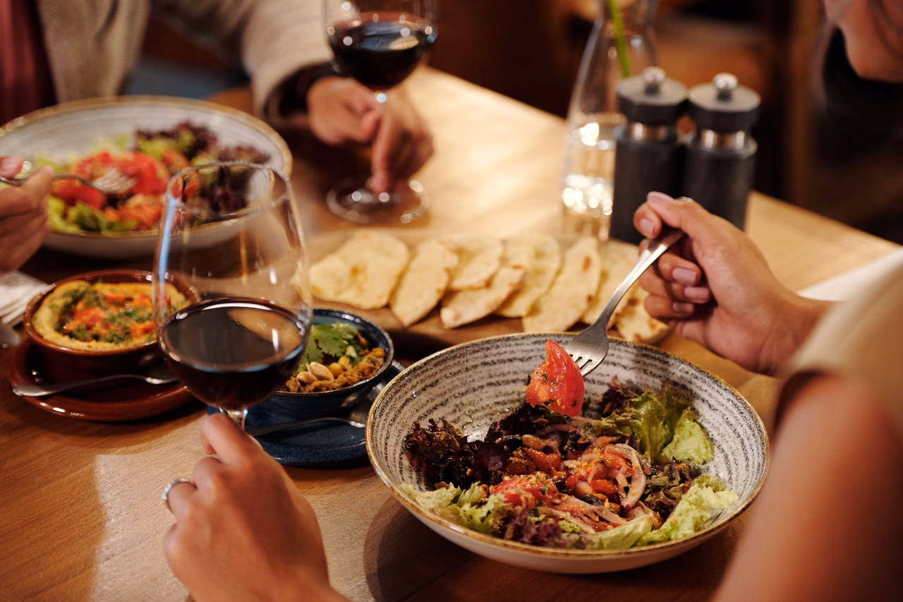 Diners share salads, hummus, flatbread and red wine across a wooden restaurant table.