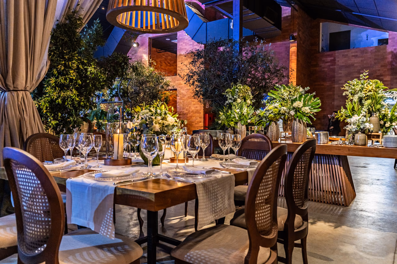 Restaurant tables set with wine glasses and white napkins among potted plants under warm pendant lights.