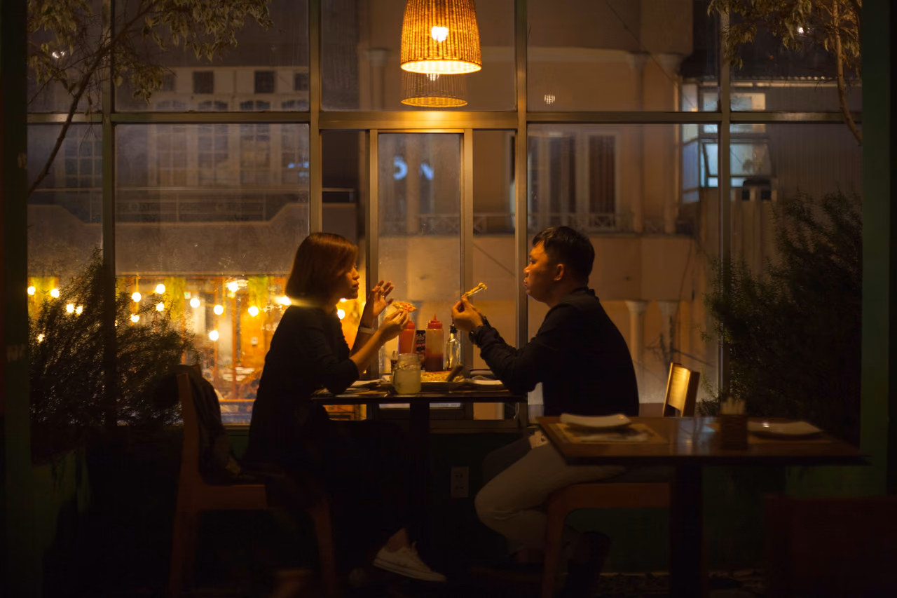 A couple eats dinner at a window table in a dimly lit restaurant under a woven pendant lamp at night.