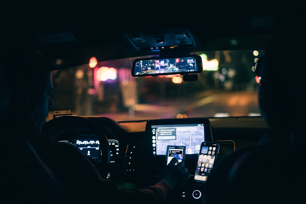 A driver taps a phone mounted on the dashboard beside a navigation screen while driving through a city at night.