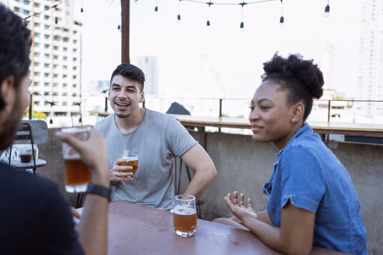 Three young friends talk over glasses of beer at a rooftop bar strung with lights, with city apartment towers behind them.