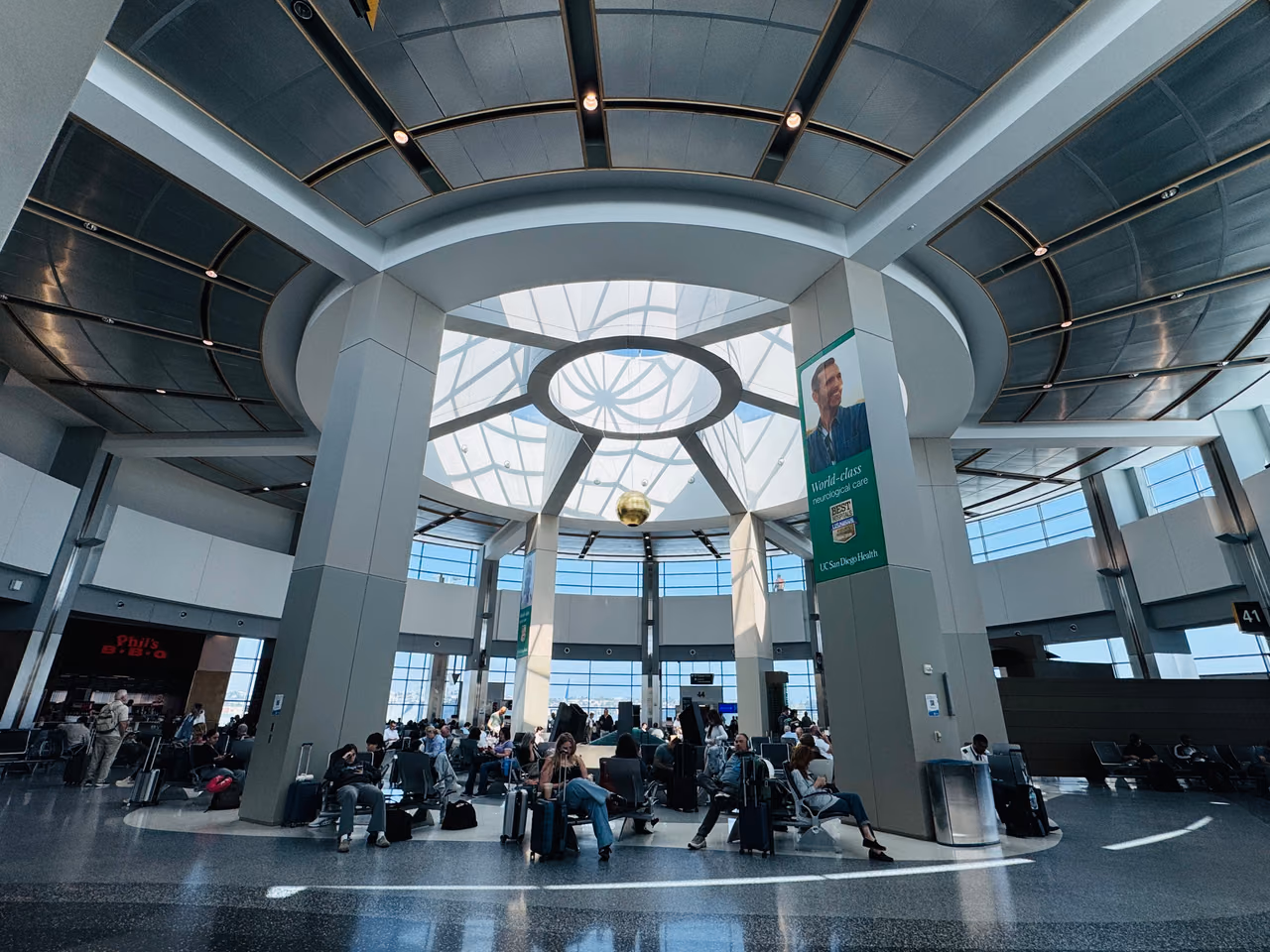 Travelers with luggage seated in a gate area beneath a domed skylight in Terminal 2 at San Diego International Airport