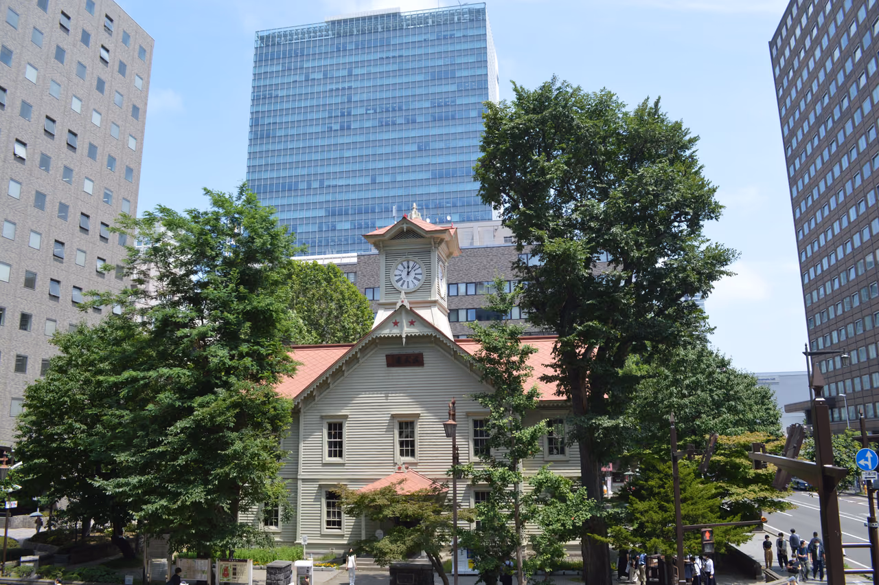 Sapporo Clock Tower, a white wooden building with a red roof, framed by green trees and office towers under a blue sky