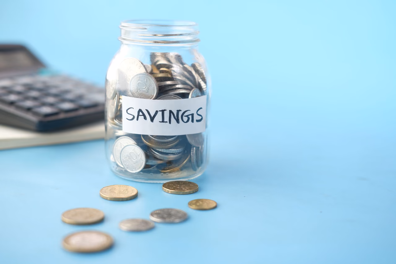 Glass jar labeled Savings filled with silver coins beside a calculator on a blue background