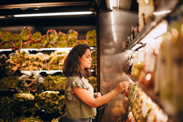 A woman in a green top reaches for packaged produce on a lit supermarket shelf, with leafy greens in the case behind her