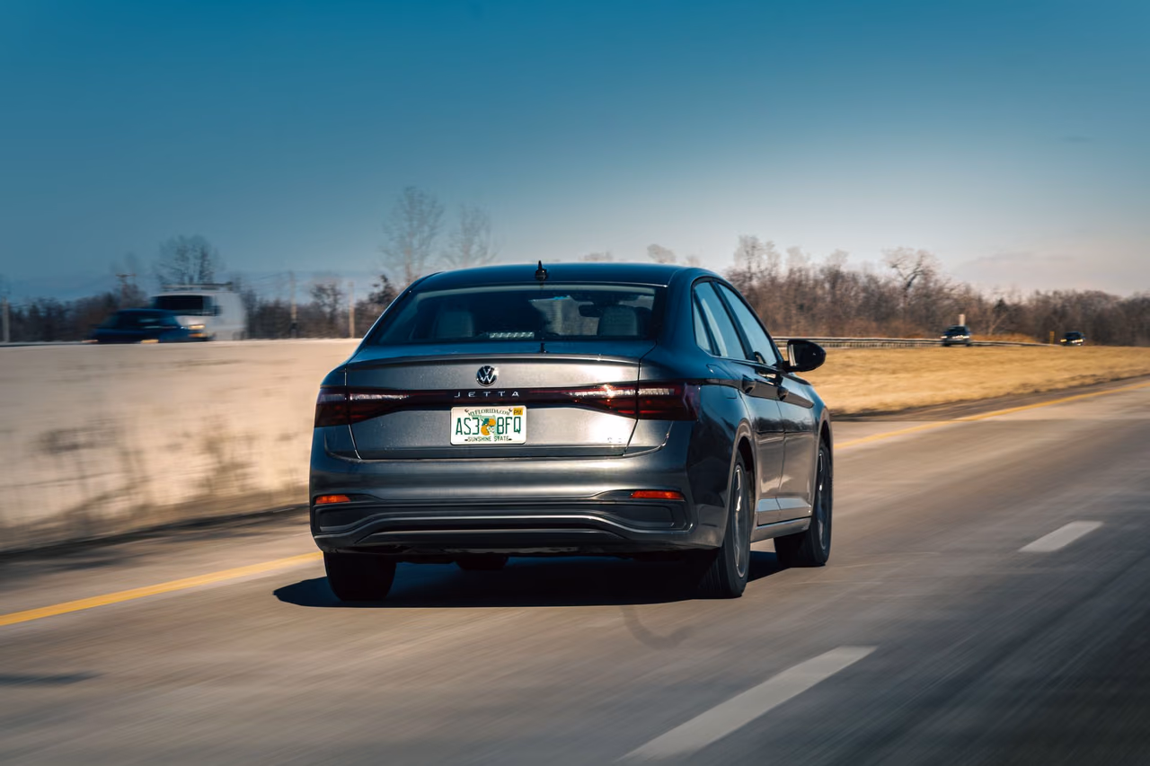 A gray sedan seen from behind driving along a highway in daylight, with bare trees and a clear sky beyond the roadside