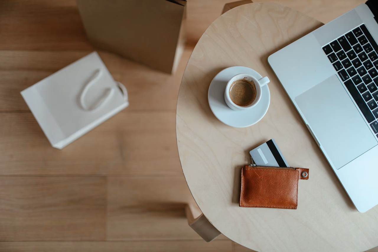 Overhead view of a single card in a small tan leather wallet on a round table beside an espresso cup and a laptop