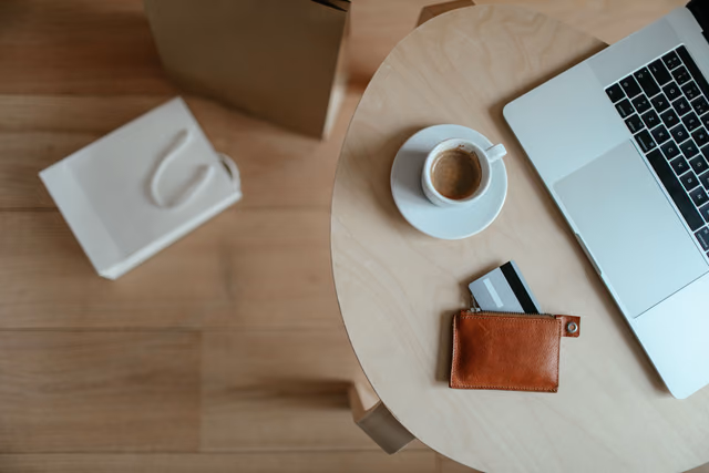 Overhead view of a single card in a small tan leather wallet on a round table beside an espresso cup and a laptop