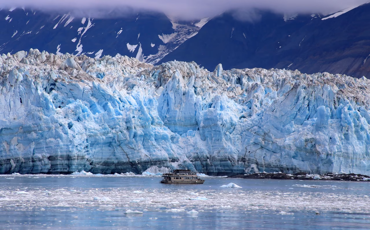 A small boat on icy water in front of a tall blue and white glacier face, with low cloud over the mountains behind it