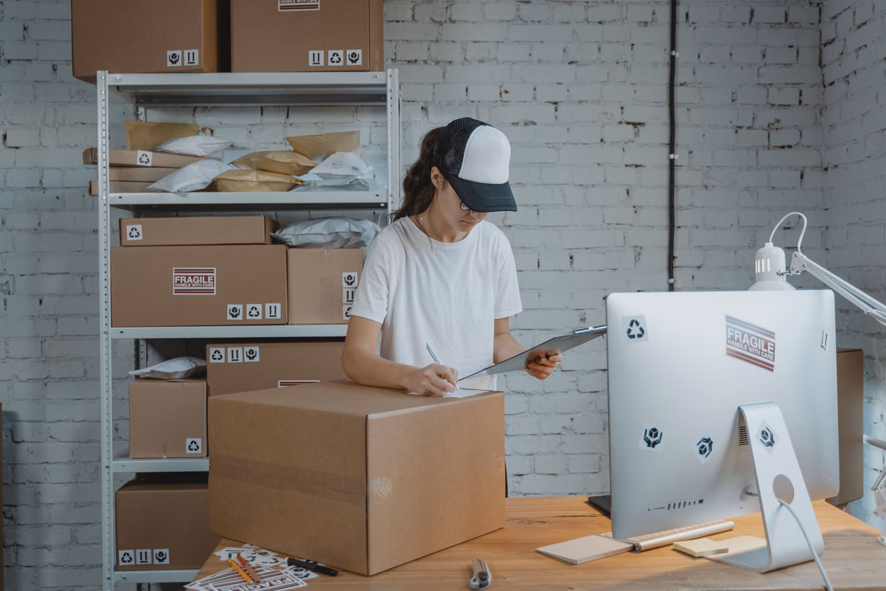 A woman in a cap checks a clipboard beside a large cardboard box, with shelves of stacked shipping boxes behind her in a small stockroom.