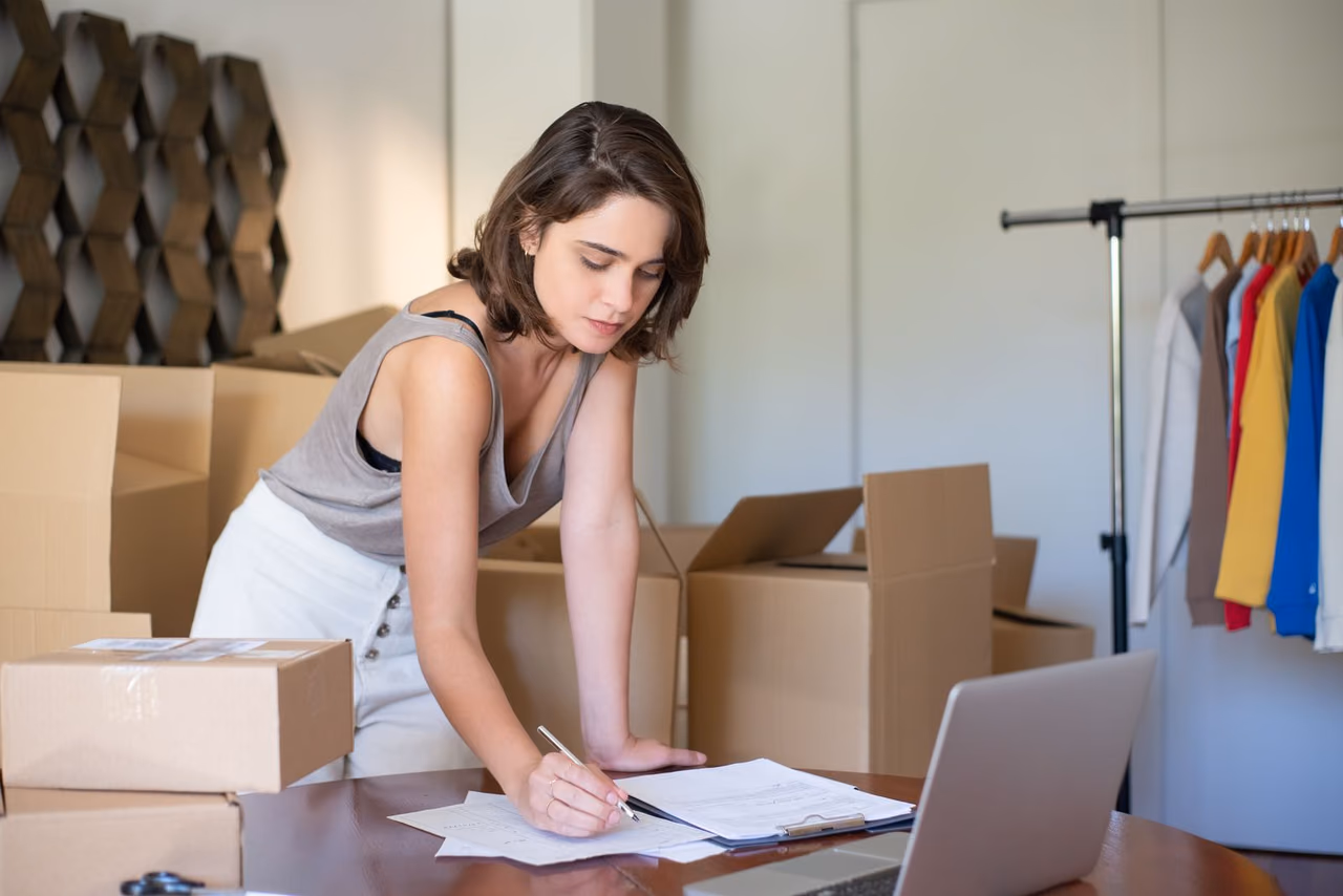 A woman leans over a desk writing on papers beside a laptop, surrounded by cardboard boxes and a rack of clothes