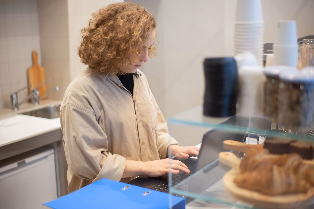 A woman in a beige shirt typing on a laptop behind a glass pastry counter in a small café