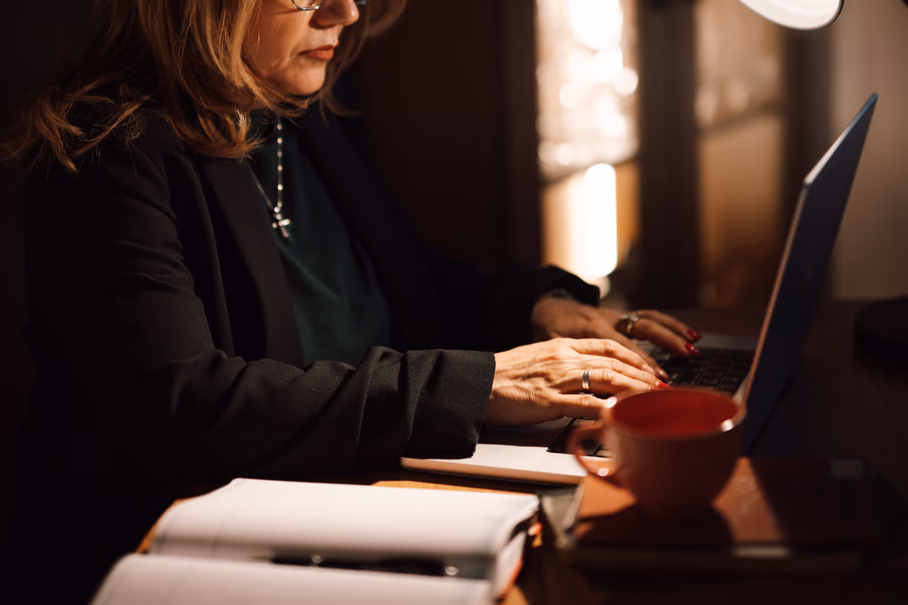 A woman in a dark blazer typing on a laptop at a wooden desk, with notebooks and a red coffee cup beside her in a dimly lit home office