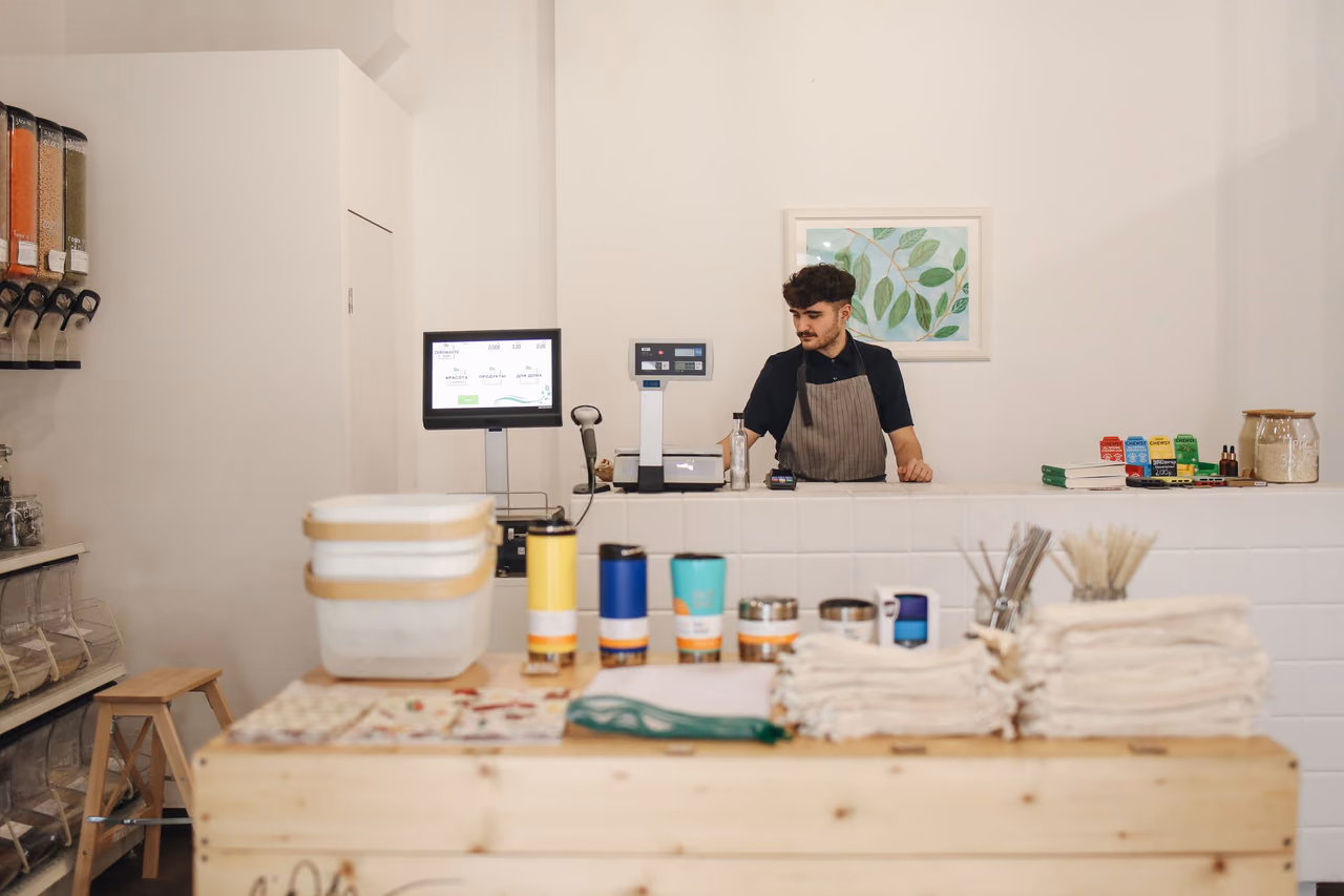 A shop worker standing behind the counter of a small refill store with stocked shelves behind him