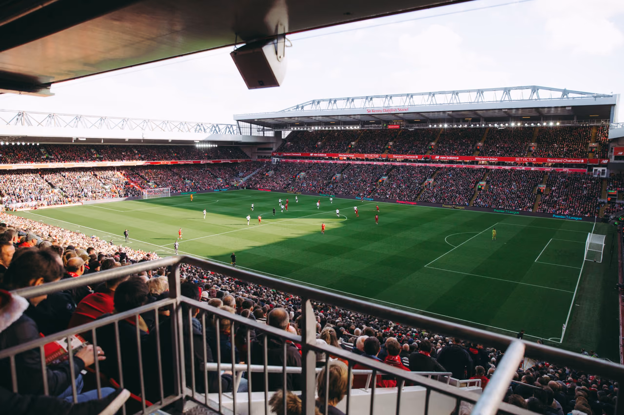 A packed soccer stadium seen from the upper stand as players contest a match on the green pitch.