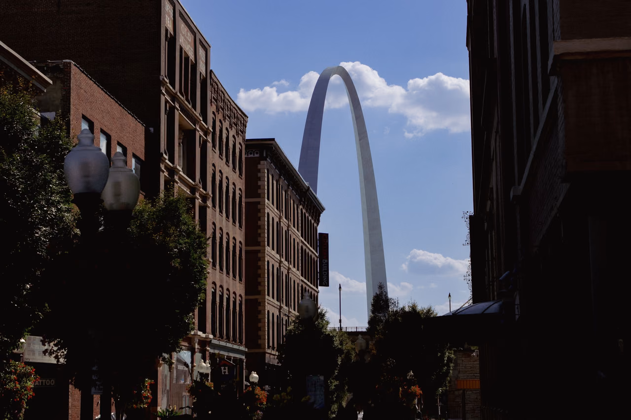 The Gateway Arch rising beyond a street of tall red brick buildings in downtown St. Louis under a blue sky