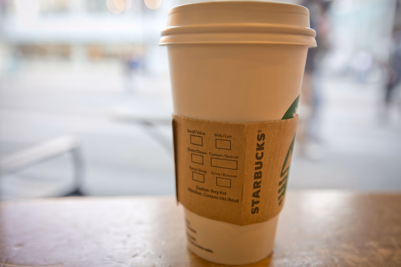 A Starbucks hot cup on a wooden window ledge, the cardboard sleeve turned to show the wordmark and the order checkboxes, with a blurred street behind