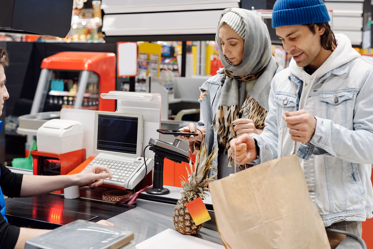 Two shoppers at a supermarket checkout lane, one paying with a phone at the card terminal while the other holds a paper grocery bag and a pineapple.