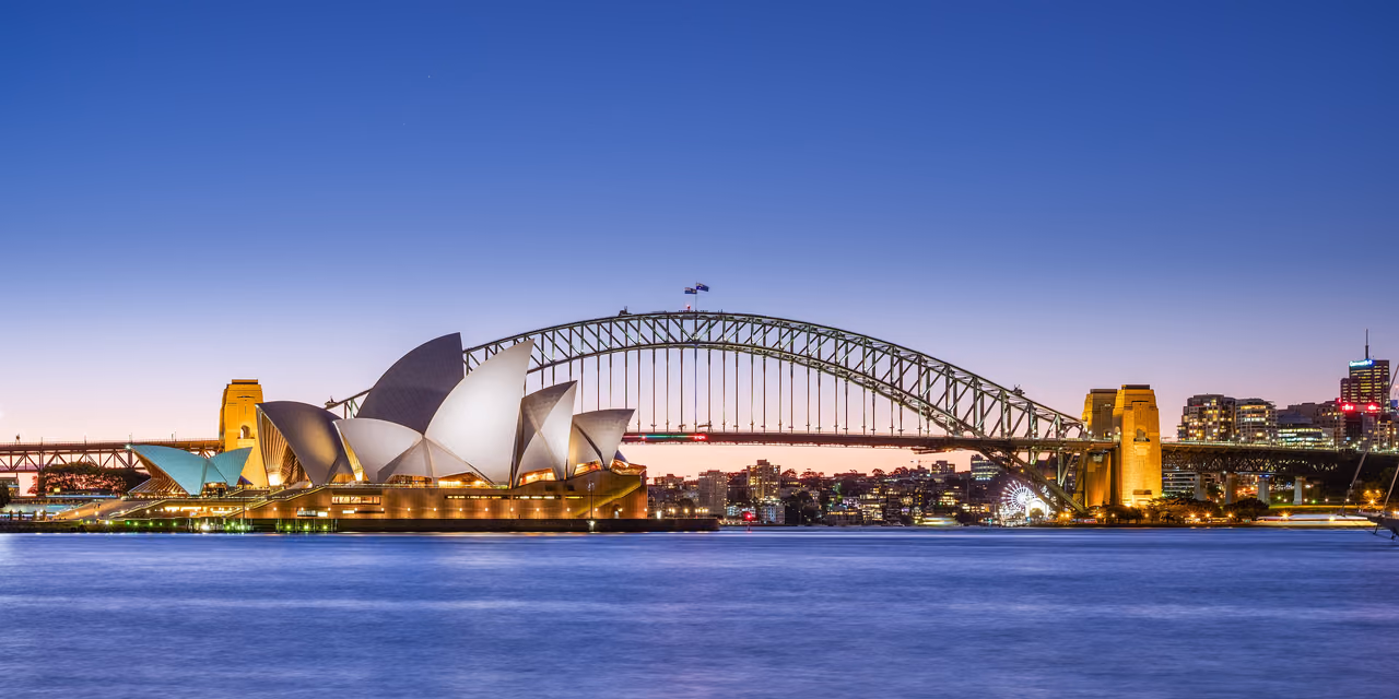 The Sydney Opera House and the Sydney Harbour Bridge at dusk, lit up across the water under a deep blue sky