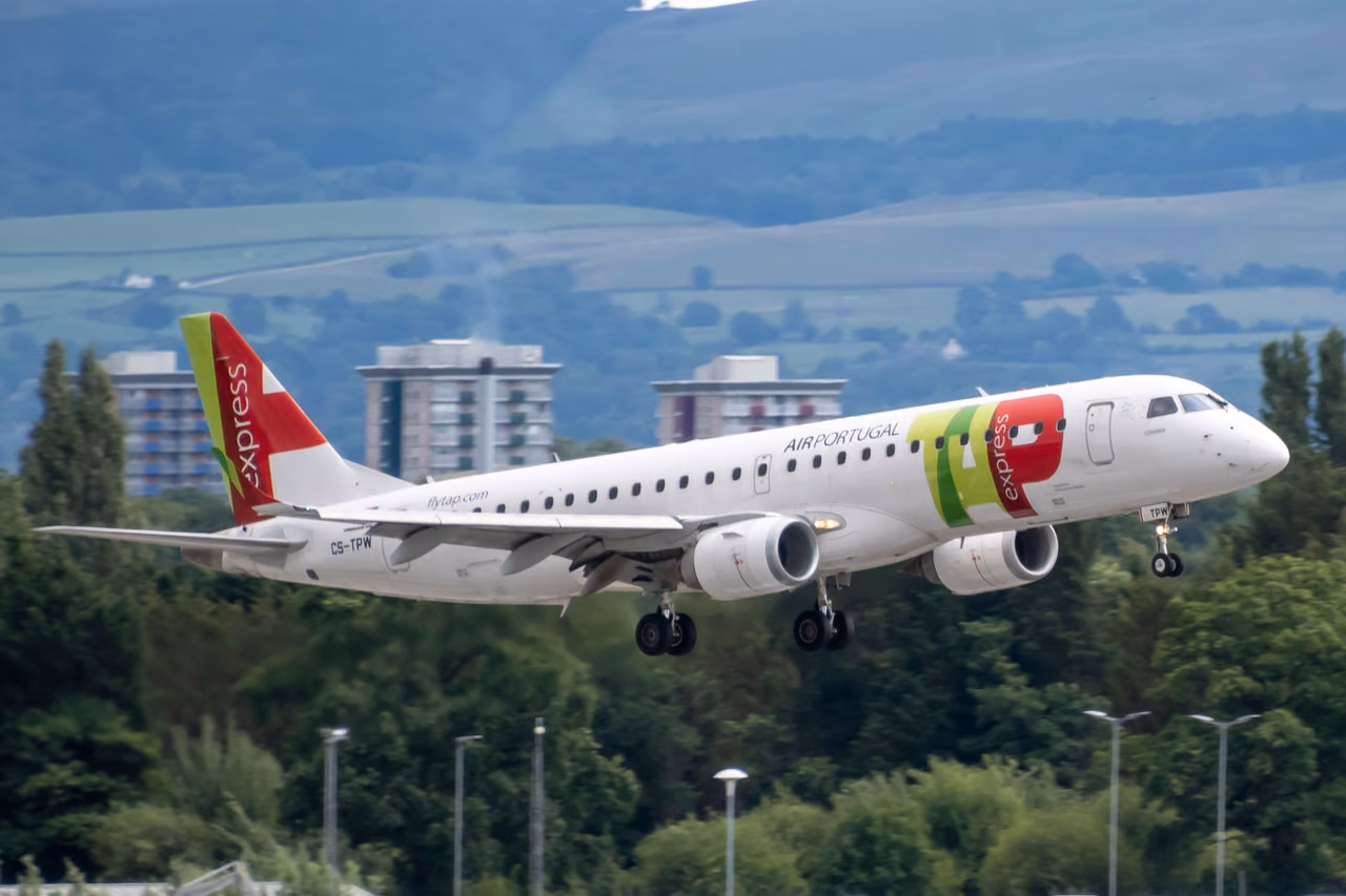 A TAP Express Embraer jet in Air Portugal colors landing over green hills, with apartment blocks behind it